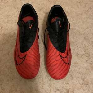 Nike röda och svarta fotbollsskor - Säljer ett par Nike fotbollsskor i rött och svart med snörning och coolt mönster. Skorna har svarta detaljer och Nike-logga på sidan samt dobbar under sulan för bra grepp på planen. Perfekta för dig som vill sticka ut på matchen!