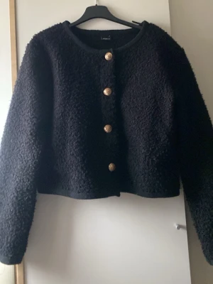 Svart teddyjacka från Gina Tricot - Mysig svart teddyjacka från Gina Tricot med fyra dekorativa guldknappar framtill. Jackan har rund halsringning, lång ärm och croppad passform. Perfekt för dig som gillar en trendig och fluffig look. Aldrig använd 