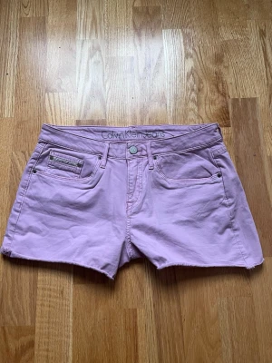 Ljusrosa shorts från Calvin Klein Jeans - Säljer ett par ljusrosa shorts från Calvin Klein Jeans. Klassisk femficksmodell med knapp och dragkedja framtill, samt bälteshällor. Tillverkade i mjuk bomull med rå kant nertill för en avslappnad vibe. Broderad detalj på bakfickorna och diskret logga på linningen.