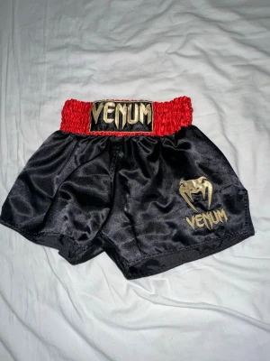 Venum svarta muay thai shorts med röd midja - Snygga svarta muay thai shorts från Venum med bred, röd resår i midjan och guldigt broderat Venum-logga framtill. Tillverkade i glansigt syntetmaterial som ger en sportig look. Perfekta för dig som gillar kampsport och vill sticka ut i ringen. Den har ett band på insidan vilket gör de möjligt att justera midjan så den sitter bra och skönt. Shortsen funkar perfekt för kampsport som Thai boxning, Boxing, Brasiliansk jiu jitsu och MMA.