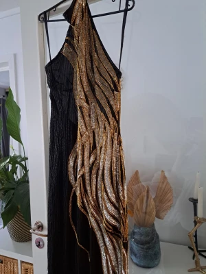 Svart långklänning med paljettdetaljer - Unik svart långklänning med glittriga paljettdetaljer i guld och silver. Klänningen har ett randigt mönster av pärlor och paljetter som ger en lyxig känsla. Perfekt för dig som vill sticka ut med en elegant och extravagant look.Tråden har släppt på.vissq ställen så bågra strass korn saknas men det syns typ inte . Klänningen var inköpt på en design second hand.