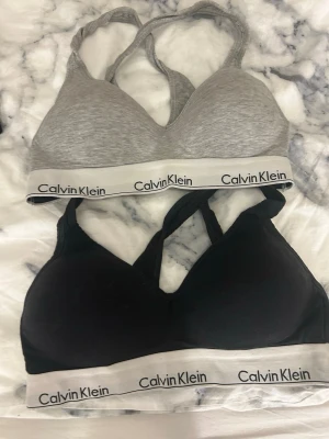 Calvin Klein sportiga toppar M - Två sportiga toppar från Calvin Klein i storlek M. En är grå och en är svart, båda med klassiskt vitt resårband med logga. De har brottarrygg och är tillverkade i mjukt bomullsmaterial. Perfekta för en chill look eller träning.