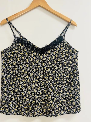 Blommig svart linnetopp med spets - Supersöt linnetopp med svart botten och små gula och lila blommor över hela. Tunna axelband och v-ringad hals med svart spetskant upptill ger en romantisk vibe. Perfekt till jeans eller kjol för en chill sommarlook.