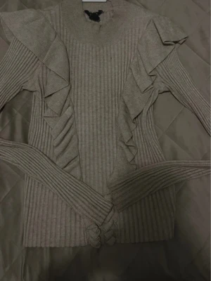 Beige ribbad tröja med volanger - Säljer en beige ribbad tröja med långa ärmar och snygga volangdetaljer längs axlar och sidor. Tröjan har en hög hals och är tillverkad i ett stretchigt stickat material som sitter snyggt på kroppen.