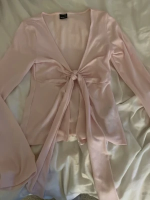 Ljusrosa omlottopp med knyt - Supersnygg ljusrosa omlottopp från Gina tricot  i ett glansigt, mjukt material. Toppen har långa ärmar, djup v-ringning och ett långt knyt framtill som ger en chic look. Perfekt att styla med jeans eller kjol för en trendig outfit.