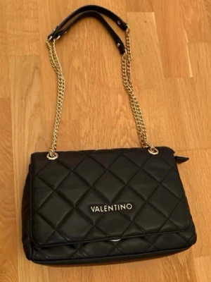 Svart quiltad axelväska från Valentino - Säljer en svart quiltad axelväska från Valentino med guldfärgade kedjor och detaljer. Väskan har ett rutmönster, lock med vridlås och axelrem i kombinerat skinn och kedja. Perfekt storlek för det viktigaste och riktigt snygg både till vardags o fest :) 