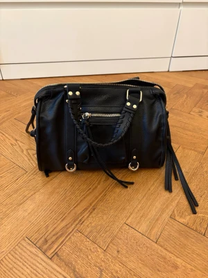 Svart handväska  - Snygg svart handväska från design by Amor skinnimitation med flätade handtag, guldfärgade metalldetaljer och dragkedja framtill. Väskan har dekorativa tofsar och flera fack. Perfekt accessoar för dig som gillar stilrena men ändå coola väskor.