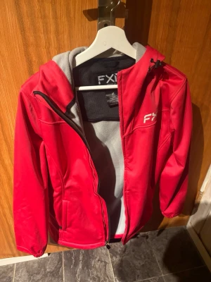 Röd vindjacka från FXR med huva - Säljer en röd vindjacka från FXR med grått innerfoder och huva. Jackan har dragkedja framtill, två sidofickor och justerbar huva med snörning. Materialet är polyester och jackan har en sportig look, perfekt för utomhusaktiviteter.