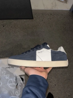 Blåvita sneakers från Pepe Jeans - Säljer ett par stilrena sneakers från Pepe Jeans i marinblått och vitt. Skorna har klassisk låg profil, rund tå och snörning. Ovandelen är i mocka och skinn, med vit sula och Pepe Jeans-logga på sidan. Perfekta till jeans eller shorts för en clean look. ORG PRIS 1200