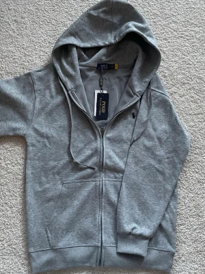 Grå Zip-Up Polo Ralph Lauren - En snygg och helt ny grå Zip-Up hoodie från Polo Ralph Launren. Den är helt ny och aldrig använd. Jag säljer den eftersom den inte passade mig.
