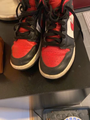 Nike Air Jordan 1 High röd/svart/vit - Säljer ett par Nike Air Jordan 1 High med klassisk färgkombination i rött, svart och vitt. Skorna har svart snörning, röd tunga med Jumpman-logga och vit sula. Tillverkade i skinn och textil, ikonisk siluett och högt skaft.