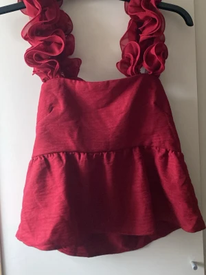 Röd volangtopp med breda axelband XS - Supersöt röd topp med breda volangprydda axelband och en lätt glansig finish. Toppen har en volangkant nedtill och knytning i ryggen för extra detalj. Perfekt för dig som vill sticka ut lite extra. Aldrig använd!!!
