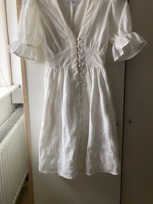 Vit kortklänning från Bubbleroom - Supersöt vit kortklänning från Bubbleroom med v-ringning, knappar framtill och volangdetaljer på ärmarna. Klänningen har ett knytband i midjan för extra fin passform och är gjord i ett lätt, strukturerat tyg. Perfekt för sommarens alla tillfällen! ENDAST ANVÄND 1gång 