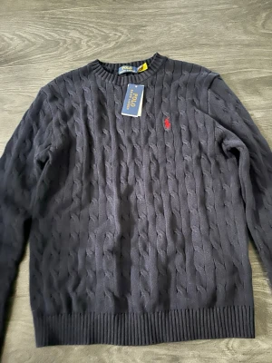 Mörkblå stickad tröja Polo Ralph Lauren - Kabelstickad mörkblå tröja från Polo Ralph Lauren med klassisk röd logga broderad på bröstet. Tröjan har rund halsringning, ribbade muddar och är långärmad. Perfekt för dig som gillar stilrena och tidlösa plagg.