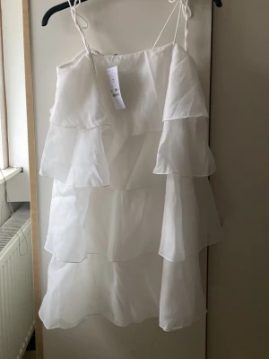 Vit volangklänning från Gina Tricot - Supersöt vit kortklänning från Gina Tricot med tunna axelband och flera lager volanger. Klänningen har en luftig och lätt känsla, perfekt för varma dagar. Materialet är tunt och ger ett flowy intryck. Modellnamn: Frilla frill mini dress. Aldrig använd 
