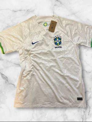 Vit Brasil Nike t-shirt med mönster - Säljer en vit Brasil t-shirt från Nike med diskret leopardmönster i tyget och blå Nike-logga på bröstet. T-shirten har korta ärmar med gröna och gula detaljer vid ärmsluten samt Brasil-märke på bröstet. Perfekt för dig som älskar fotboll och vill sticka ut.