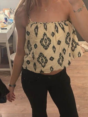 Beige croppad linnetopp med mönster - Säljer en beige croppad linnetopp med svarta grafiska mönster. Toppen har tunna axelband och en luftig passform som ger en avslappnad vibe. Perfekt till sommaren och enkel att styla med jeans eller kjol.