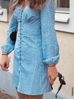 Blå blommig kortklänning med knappar - Supersöt blå kortklänning med små vita blommor, långa puffärmar och knappar hela vägen framtill. Klänningen har en feminin passform och v-ringning, perfekt för vår och sommar. Lätt och skönt material som känns luftigt.