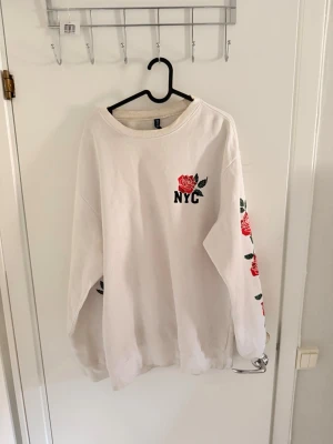 Vit sweatshirt med rosor och NYC-tryck - Vit oversized sweatshirt från H&M med broderade röda rosor på ena ärmen och ett NYC-tryck med ros på bröstet. Tröjan har rund halsringning och långa ärmar, perfekt för en chill och trendig look.