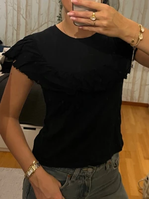 Svart topp från Zara - Supersnygg svart topp från Zara med volangdetaljer över axlar och bröst. Toppfn har rund halsringning och är kortärmad. Perfekt att styla med jeans eller kjol för en trendig look. Passar xs-m