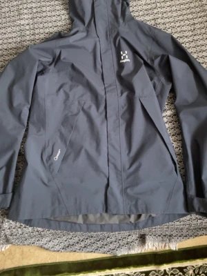 Mörkgrå vindjacka från Haglöfs dam M - Snygg mörkgrå vindjacka från Haglöfs för dam i storlek M. Jackan har huva, dragkedja framtill och två snedställda fickor. Tillverkad i slitstark polyester och perfekt för blåsiga dagar. Enkel och stilren design med diskret logga på bröstet.