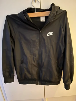 Svart vindjacka från Nike - Säljer en svart vindjacka från Nike med huva och dragkedja framtill.  Storleken på jackan är 150-158cm. Jackans vänsterficka har dock en dragkedja som är sönder den går fortfarande att stänga men det är lite svårt.