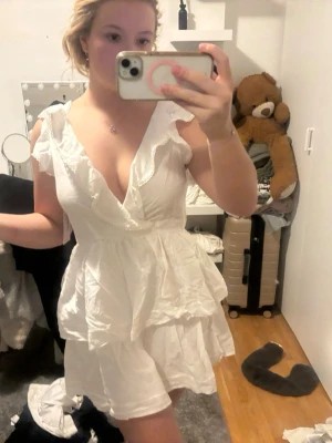 Vit volangklänning från Nelly XS - Supersöt vit kortklänning från Nelly med volanger, djup v-ringning och öppen rygg med knyt. Klänningen har ärmlös design och omlott framtill, perfekt för sommarens alla tillfällen. Lätt och luftig känsla med fina spetsdetaljer längs kanterna.