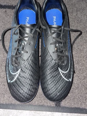 Nike Phantom svart/blå fotbollsskor - Säljer ett par Nike Phantom fotbollsskor i svart med blå detaljer och vit swoosh. Skorna har snörning, rund tå och platt sula med dobbar för bra grepp på planen. Ovandelen är i syntetmaterial med textur för bättre bollkontroll. Perfekta för dig som vill ta spelet till nästa nivå.