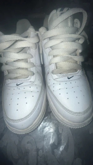 Nike Air Force 1 vita sneakers - Säljer ett par klassiska Nike Air Force 1 sneakers i helvitt skinn med vit sula och snörning. Skorna har den ikoniska swoosh-loggan på sidan och AIR-detalj på hälen. Perforerade detaljer på tån för extra ventilation och stilren look. Perfekta för dig som gillar tidlös streetwear.