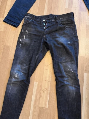 Svarta Dsquared2 jeans med slitningar - Snygga svarta jeans från Dsquared2 med coola slitningar och tvättade detaljer. Klassisk femficksmodell, knappgylf och Dsquared2-logga bak i midjan. Jeansen har en rak passform och är tillverkade i bomull. Perfekt för dig som gillar streetstyle och vill sticka ut.