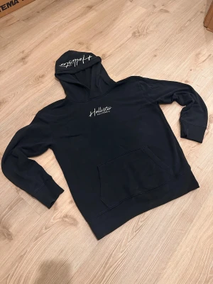 Svart hoodie från Hollister - Svart hoodie från Hollister med vit logga på bröstet och huvan. Klassisk känguruficka framtill och ribbade muddar vid ärmslut och nederkant. Perfekt för en chill och avslappnad stil.