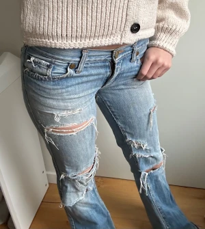 Ljusblå slitna bootcut jeans - Säljer ett par ljusblå jeans med bootcut och snygga slitna detaljer över benen. Jeansen har låg midja och klassiska fickor, samt broderad bakficka med guldfärgad tråd. Perfekta för en avslappnad och edgy stil.