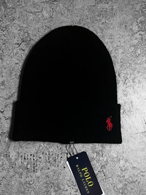 Svart mössa från Polo Ralph Lauren - Svart ribbstickad mössa från Polo Ralph Lauren med klassisk uppvikt kant. Märkets ikoniska röda broderade logga med polospelare på framsidan. Mjuk och stretchig kvalitet, perfekt för kalla dagar. Snygg och enkel design som funkar till allt.