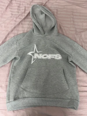 Grå hoodie från NOFS med tryck - Snygg grå hoodie från NOFS med stort vitt tryck på bröstet och en stjärndetalj. Tröjan har huva, magficka och är fodrad med mjuk fleece på insidan. Perfekt för chill dagar och streetstyle. Långärmad och med ribbade muddar för extra komfort.