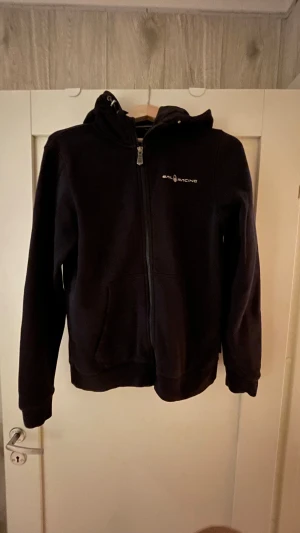 Svart huvtröja från Sail Racing - Svart hoodie från Sail Racing med dragkedja och huva. Tröjan har två fickor framtill och en snygg logga på bröstet samt en detalj på huvan.