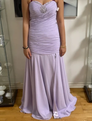 Ljuslila långklänning med strass - Säljer en elegant ljuslila långklänning med v-ringad överdel, draperad midja och glittriga strassdetaljer vid bysten. Klänningen har smala axelband och ett svepande, utsvängt kjolparti som ger en riktigt drömmig siluett. Perfekt för bal eller student!