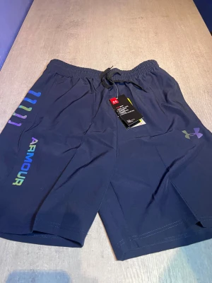 Blå träningsshorts från Under Armour - Snygga marinblå träningsshorts från Under Armour med elastisk midja och snörning. Shortsen har färgglada logotyper på benet och är gjorda i ett lätt, snabbtorkande syntetmaterial som andas. Perfekta för gymmet eller löprundan.