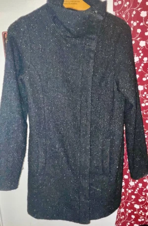 Svart kappa från Vero Moda - Stilren svart kappa från Vero Moda med diskret glittrig yta. Jackan har hög krage, dold knäppning och två fickor framtill. Materialet är grovt vävt och fodret är slätt och svart. Perfekt för dig som gillar en klassisk look med en twist.