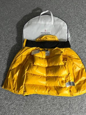 Svart och gul Moncler dunjacka - Säljer en fet svart och gul dunjacka från Moncler med coola detaljer. Jackan har en gul insida, svart utsida, dragkedjor med snören och Moncler-logga på ärmen. Puffig modell med huva och schyssta fickor. Perfekt för dig som vill sticka ut i vinterkylan. STORLEKEN ÄR 1 INOM MONKLER
