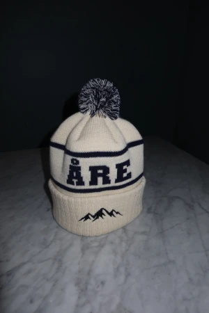 Vit Åre mössa  - Riktigt fräsch Åre mössa från SkiStyle UF✅. Framsidan har ÅRE-text och ett broderat bergmotiv och färg cream. Stickad i mjukt material, otrolig kvalitet och perfekt för kalla dagar, skriv vid frågor eller funderingar!