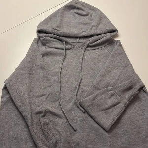 Grå hoodie med snörning - En enkel och chill grå hoodie med huva och snörning. Tröjan har långa ärmar med ribbade muddar och är gjord i mjukt bomullsmaterial. Perfekt för att slänga på sig när man vill vara bekväm och avslappnad.