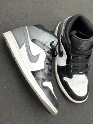 Nike Air Jordan 1 svart/vit/grå sneakers - Säljer ett par Nike Air Jordan 1 sneakers i svart och vitt skinn med klassisk high-top design. Skorna har svarta snören, perforerad tå och ikoniska Jordan-loggor på sidan och plösen. Perfekta för dig som gillar streetstyle och vill ha en tidlös look.
