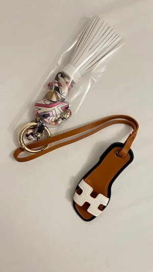 Färgglad nyckelring med tofs och scarf - Säljer en unik accessoar med en vit lädertofs, flätad scarf i rosa, beige, svart och guld med grafiskt mönster, samt en brun och svart läderdetalj med vit dekor. Allt sitter på en guldfärgad ring. Perfekt att hänga på väskan eller använda som nyckelring.