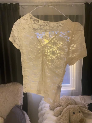 Vit spetsblus med omlott - Säljer en söt vit blus i spets med omlott-design och korta ärmar. Blusen har v-ringning och är genombruten med blommigt spetsmönster, perfekt för en romantisk look. Passar dig som gillar ljusa och feminina plagg.