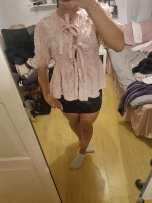 Ljusrosa blus med knyt och struktur - Supersöt ljusrosa blus med strukturmönster och två knyt framtill. Blusen har puffiga trekvartsärmar och lätt volang nertill. 