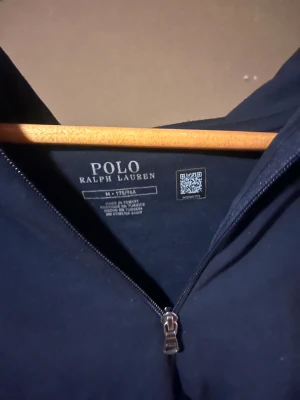 Mörkblå hoodie från Polo Ralph Lauren - Säljer en mörkblå hoodie från Polo Ralph Lauren med dragkedja och huva. Tröjan har långa ärmar, snörning vid huvan och en vit broderad logga på bröstet. Materialet är mjuk bomull och den har en klassisk känguruficka framtill.