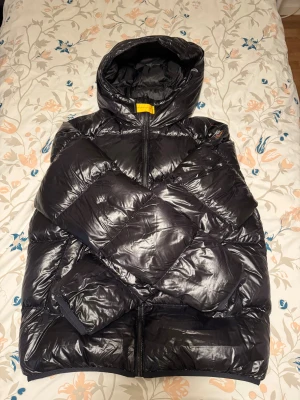 Svart pufferjacka från Parajumpers - Säljer en svart pufferjacka från Parajumpers, modell Buzz Puffers. Jackan har en glansig finish, huva och dragkedja framtill. Insidan har en meshficka med orange kant. Perfekt för kalla dagar och har en modern, oversized vibe. Den passar för s-xs