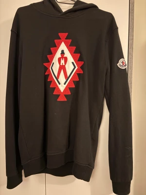 Svart hoodie från Moncler med broderi - Svart hoodie från Moncler i 100% bomull med ett stort broderat märke i rött, vitt och svart på bröstet. Klassisk huva och lång ärm, samt Moncler-logga på ena ärmen. Perfekt för dig som gillar streetwear med exklusiv känsla.