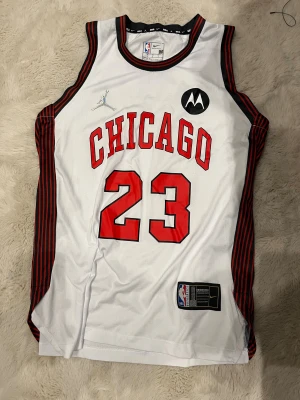 Chicago Bulls Jordan 23 fotbollströja - Vit fotbollströja med svarta och röda detaljer, Chicago och nummer 23 i rött framtill. Sidorna har svarta och röda ränder. Materialet är mesh för bra andningsförmåga. Silverfärgad logga på framsidan och klassisk Jordan-stil.