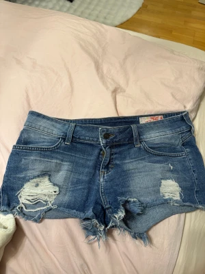 Lågmidjade shorts  - Snygga blå jeansshorts med slitna detaljer och fransig kant. Framtill har de coola hål och bakfickorna är broderade med ett unikt mönster. Klassisk femficksmodell och knappstängning. Perfekt för dig som gillar streetstyle och vill sticka ut. Pris går att diskutera💕